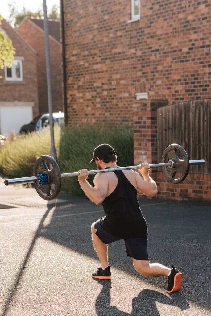 Back view of anonymous strong male athlete in sports clothes doing walking lunges with barbell on asphalt during training